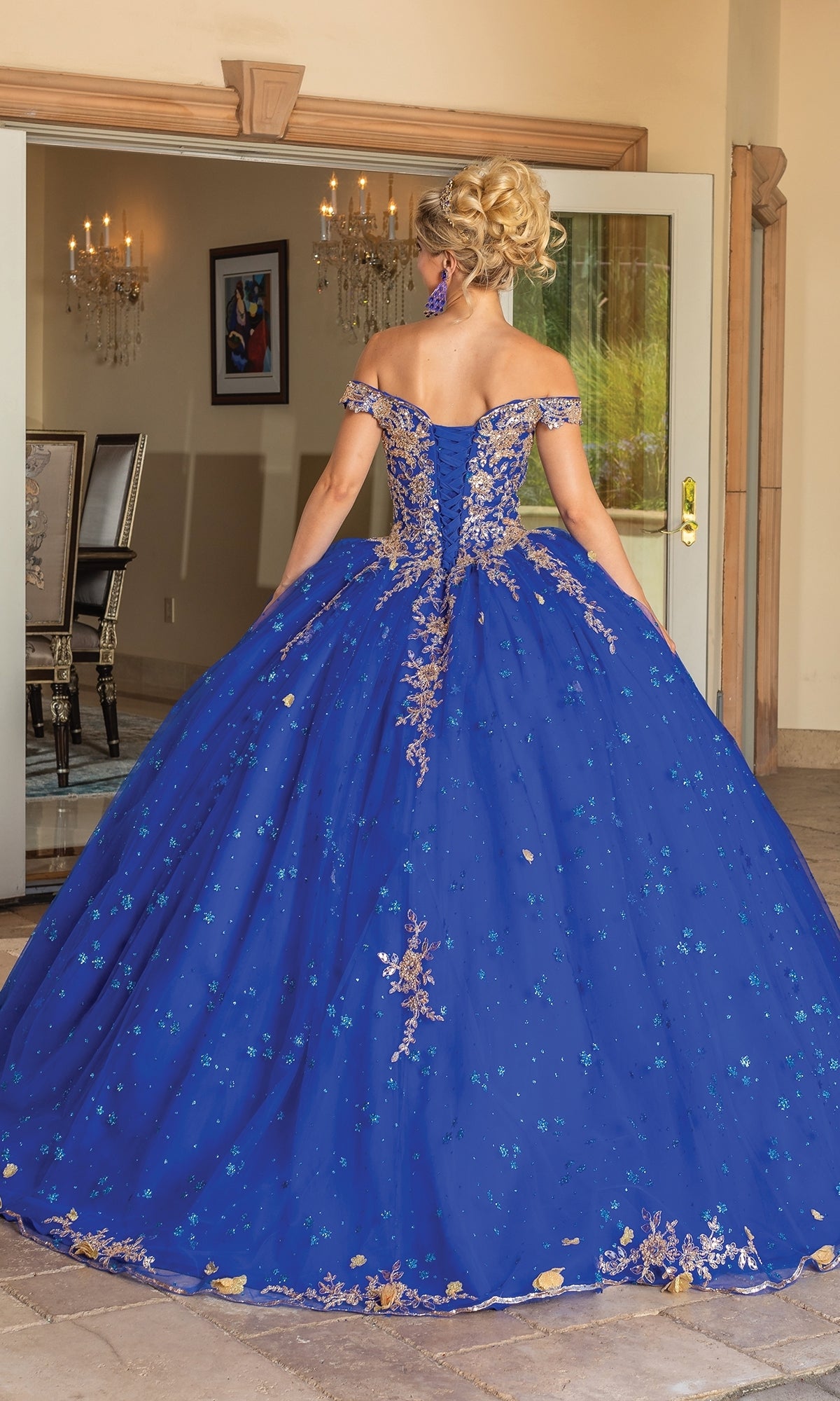 A blonde woman with an updo stands indoors with her back to the camera, wearing Quinceanera Dress 1796 By Dancing Queen—a blue off-the-shoulder gown with gold embellishments.